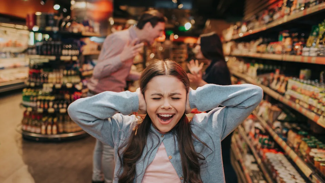 A child covering her ears while her parents argue behind her in a store aisle.